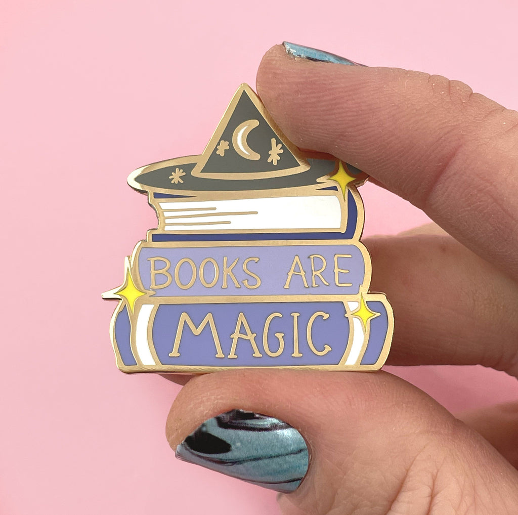 A hard enamel lapel pin being held in a hand. The pin is purple and white and says Books are Magic with books and a witch’s hat.  