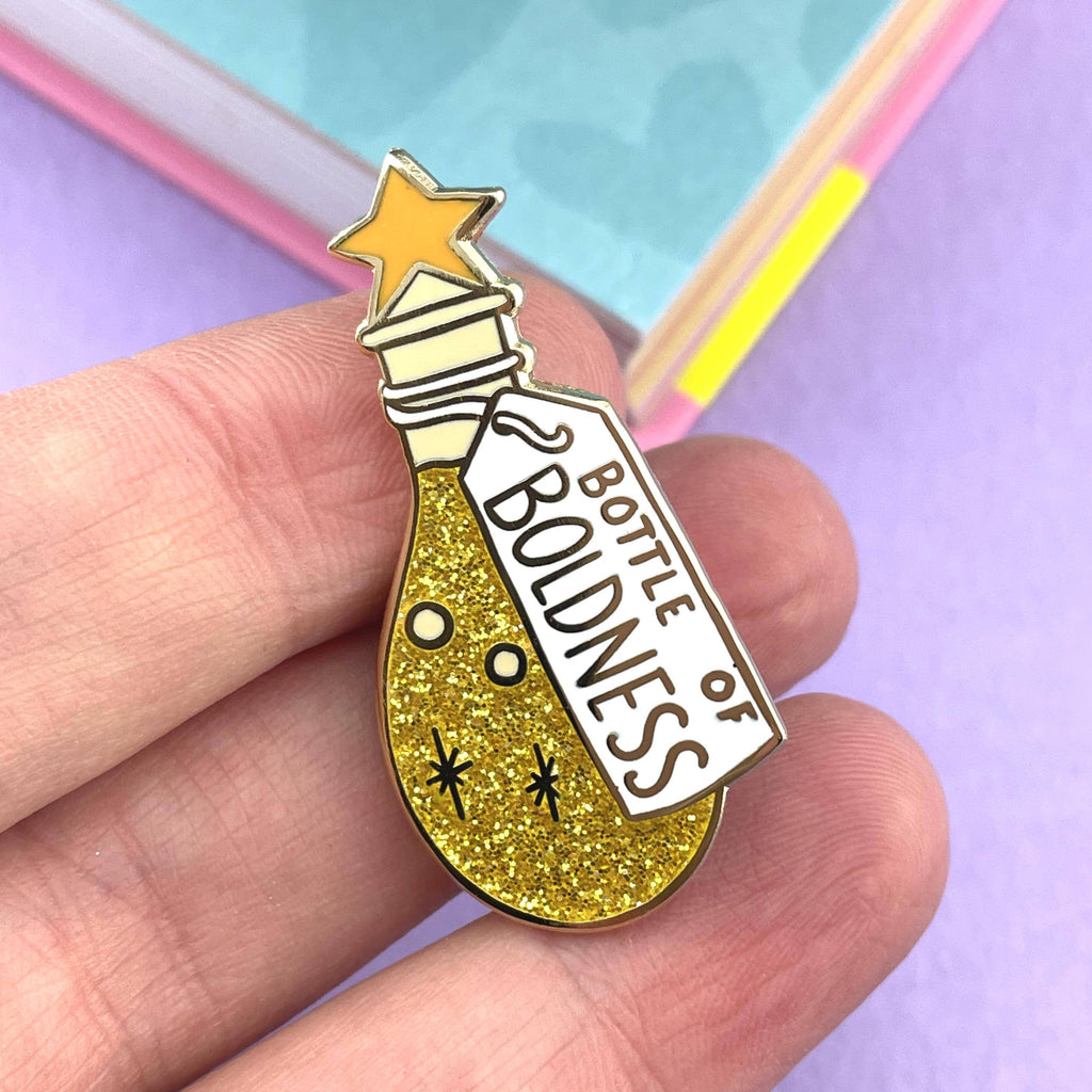 A hard enamel lapel pin being held in a hand. The pin is in the profile of a bottle with yellow glitter. The pin says Bottle of Boldness.
