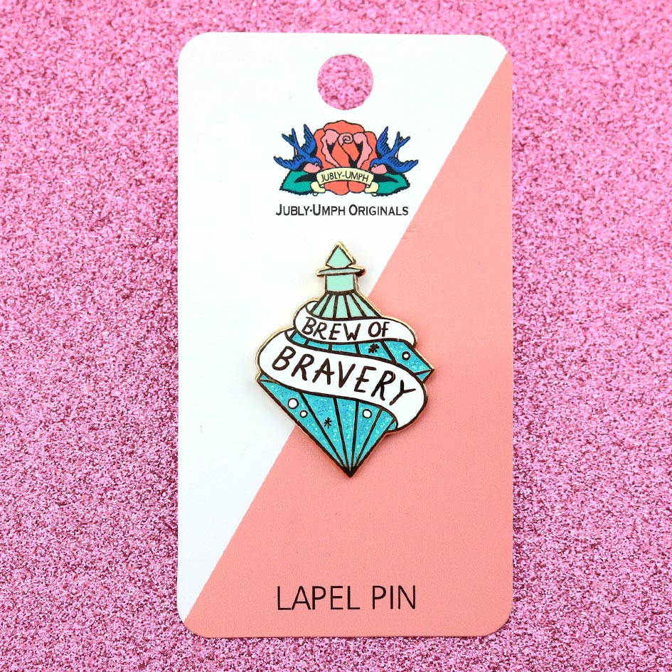 A hard enamel lapel pin on Jubly-Umph cardstock. The pin is in the profile of a diamond shaped bottle with blue glitter. The pin says Brew Of Bravery.