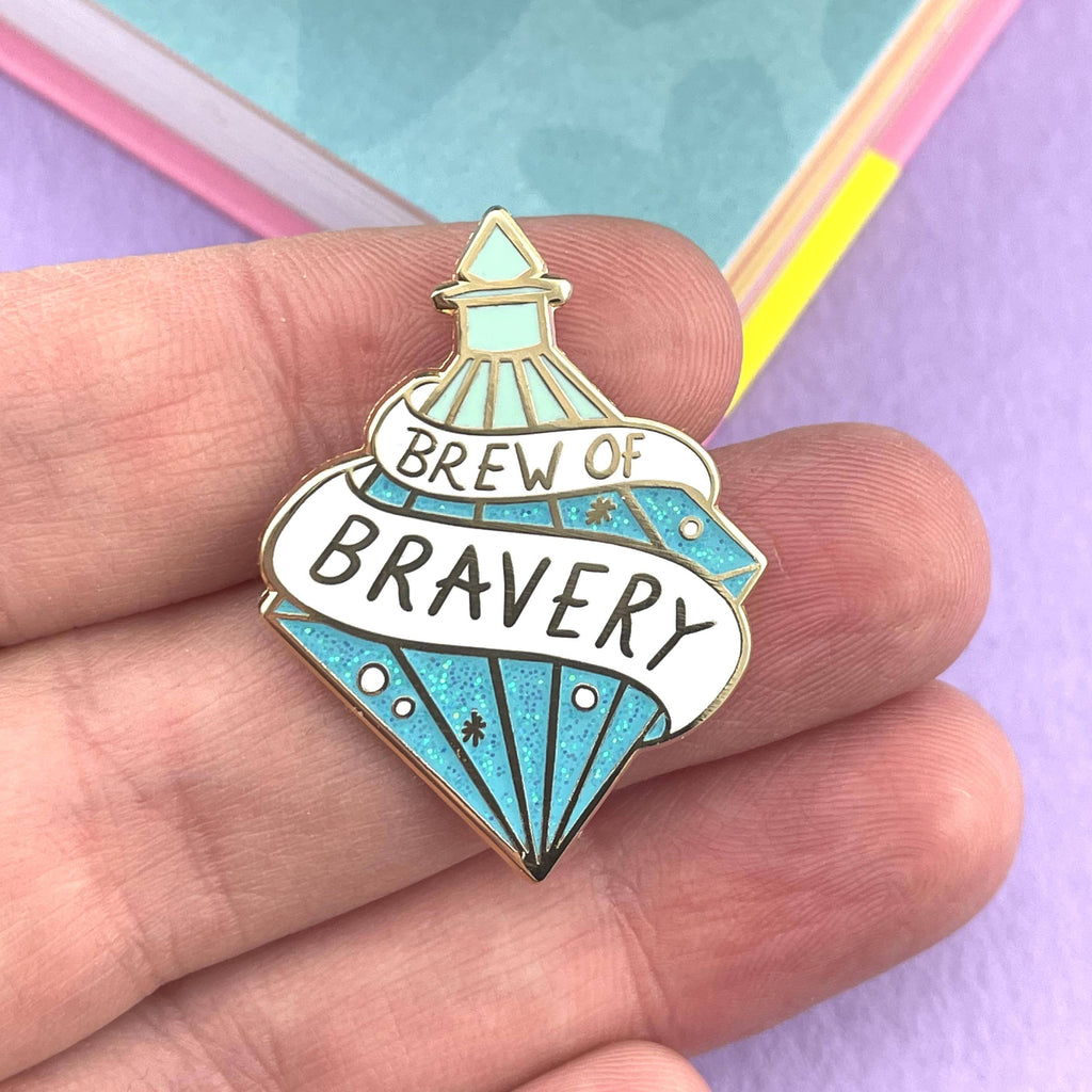 A hard enamel lapel pin being held in a hand. The pin is in the profile of a diamond shaped bottle with blue glitter. The pin says Brew Of Bravery.