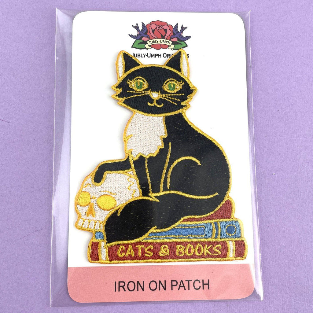 An iron on embroidered patch on Jubly-Umph cardstock against a purple background. The patch is a black and white cat with a scull sitting on books. The patch reads Cat’s and Books.