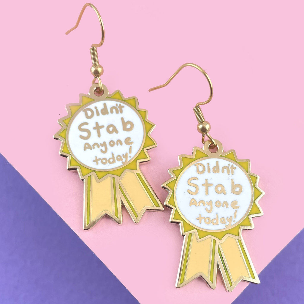 A pair of dangle earrings in the shape of an award ribbon. They are on a pink and purple background. The ribbon is yellow and white, and reads Didn’t Stab Anyone Today!