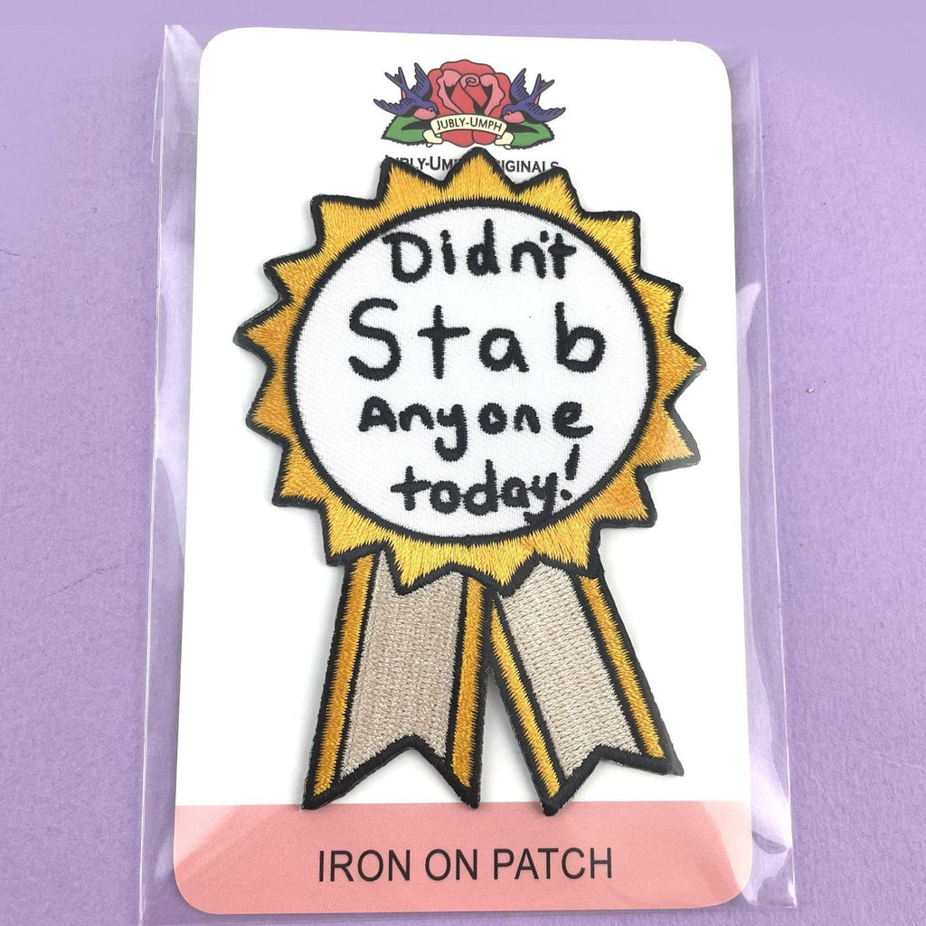 An iron on embroidered patch on Jubly-Umph cardstock. The patch is in the shape of an award ribbon. The ribbon is yellow and white, and reads Didn’t Stab Anyone Today!