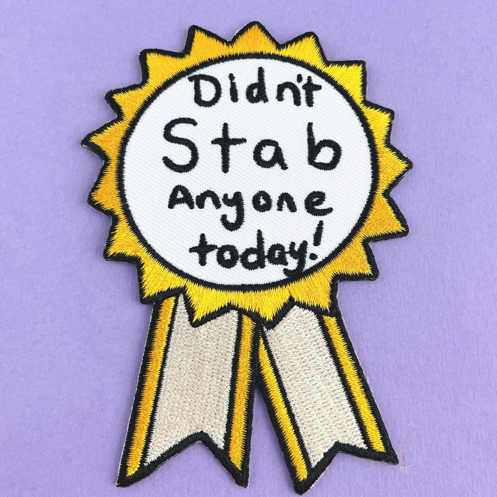 An iron on embroidered patch on purple background. The patch is in the shape of an award ribbon. The ribbon is yellow and white, and reads Didn’t Stab Anyone Today!