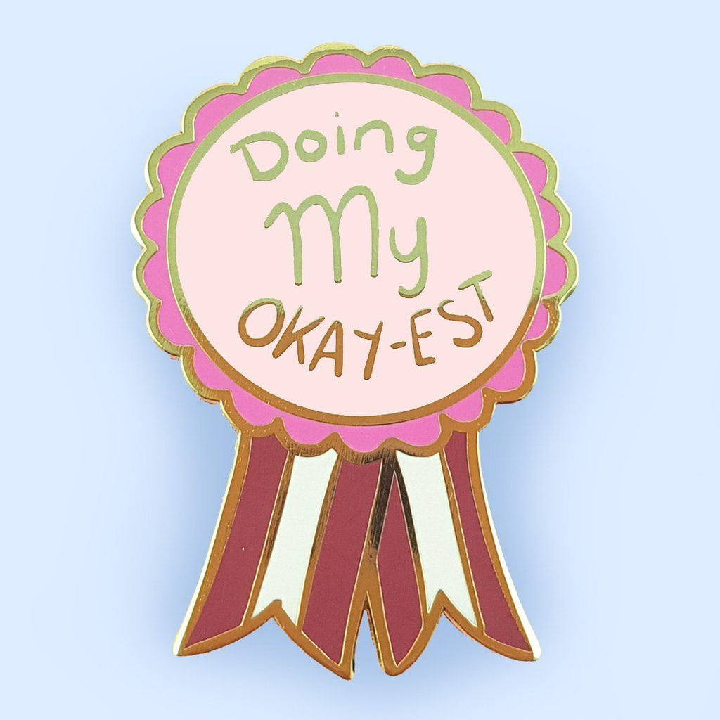 A hard enamel lapel pin on a blue background. The pin is in the shape of an award ribbon. The ribbon is dark pink and light pink and reads Doing my Okay-est.