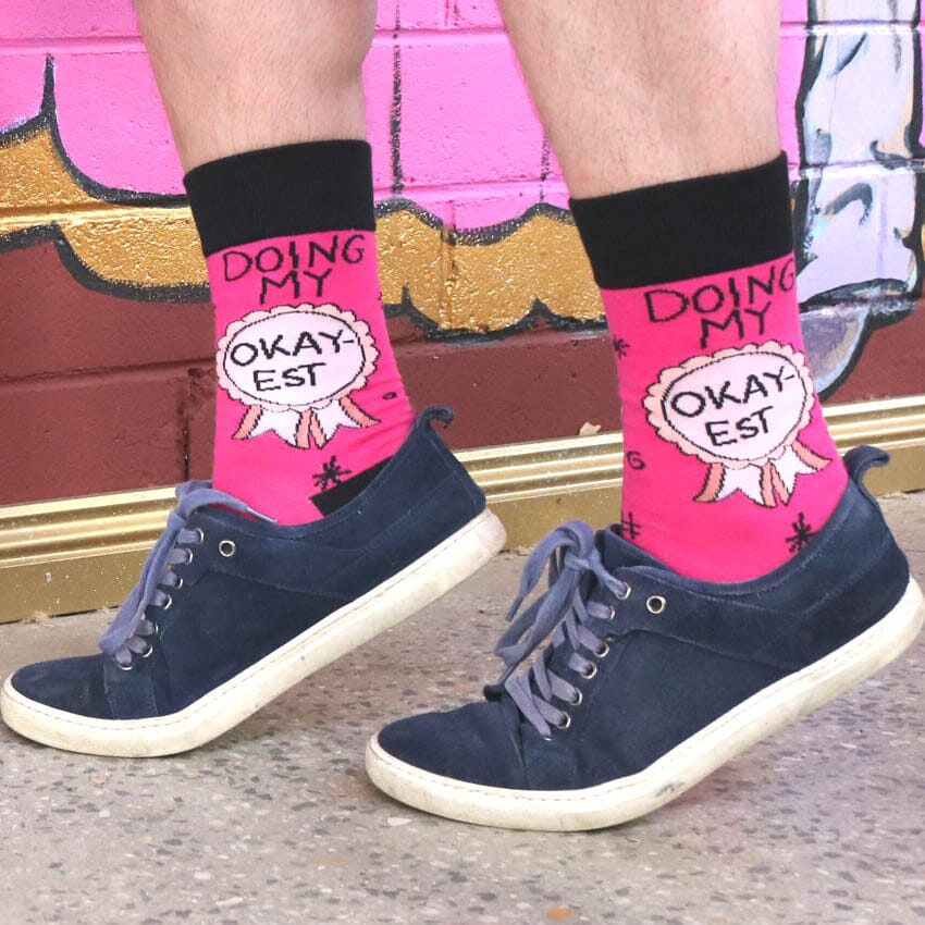 A pair of socks worn with blue shoes. The socks are pink and black and read Doing My Okay-est.