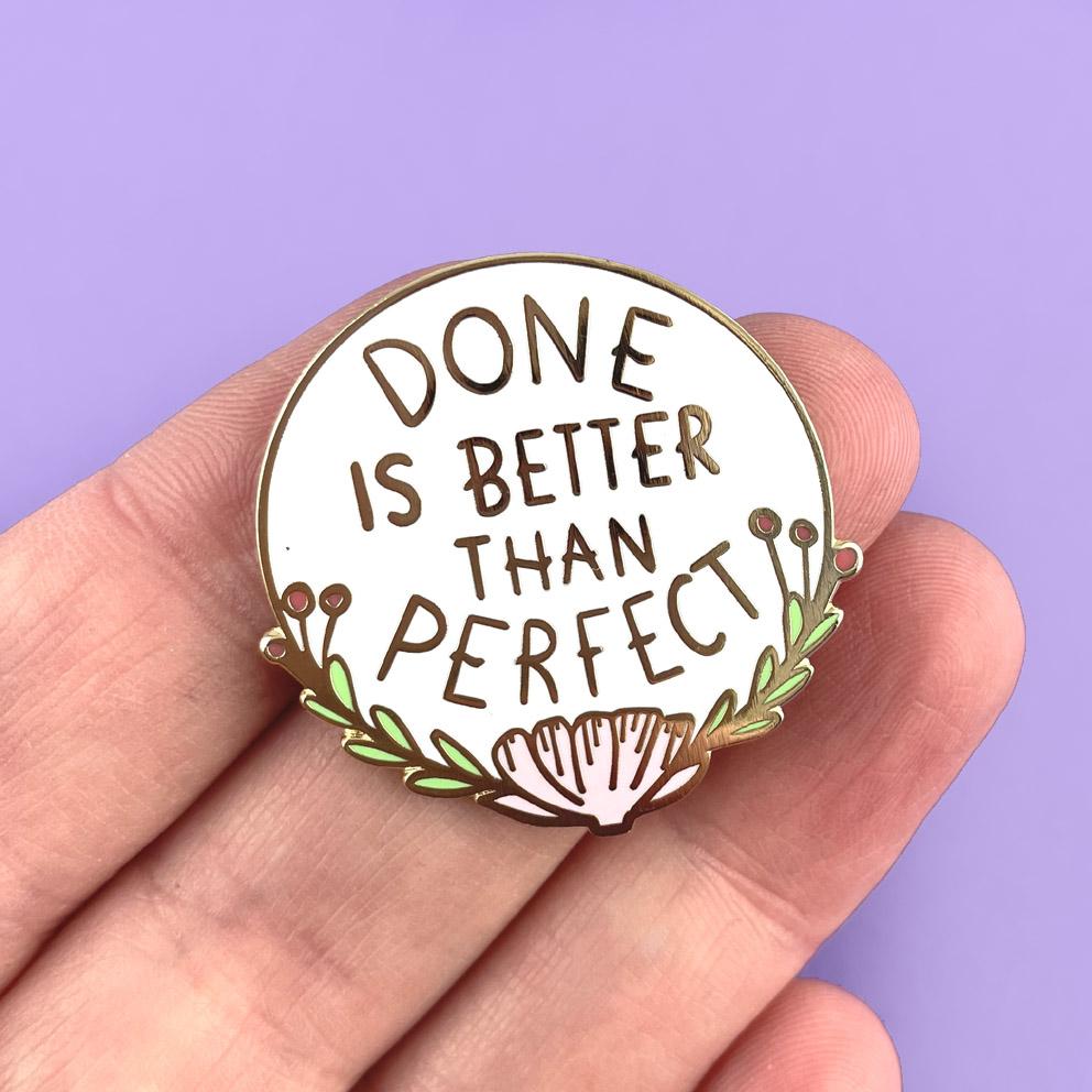 A hard enamel lapel pin being held in a hand. The pin is white with gold lettering and reads Done Is Better Thank Perfect.