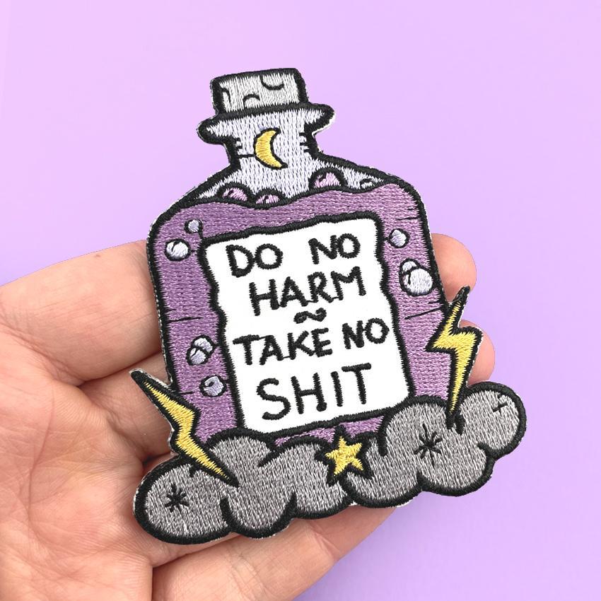 An iron on embroidered patch being held in hand on purple background. The patch is purple and in the shape of a bottle with clouds and lightning bolts. The pin reads Do No Harm Take No Shit.