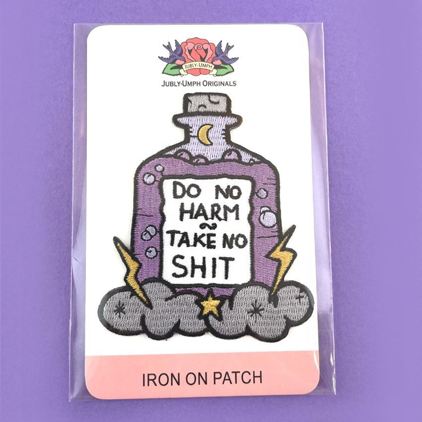 An iron on embroidered patch on Jubly-Umph cardstock. The patch is purple and in the shape of a bottle with clouds and lightning bolts. The pin reads Do No Harm Take No Shit.