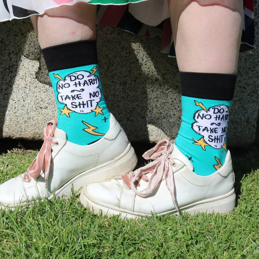 A pair of socks worn with white shoes. The socks are teal and black and read Do No Harm Take No Shit.