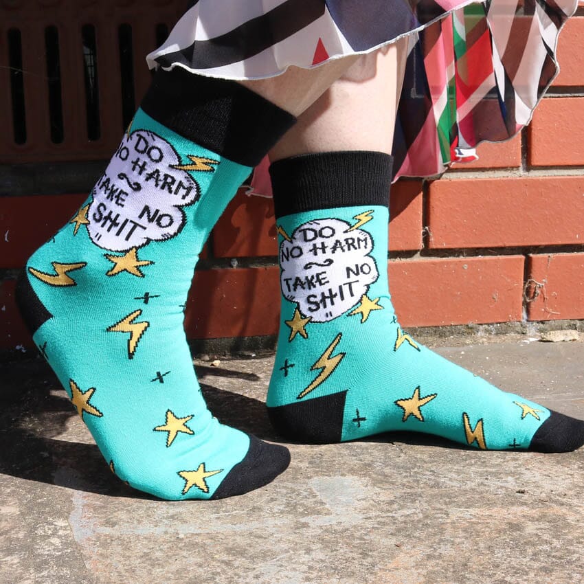 A pair of socks being modelled against a brick wall. The socks are teal and black and read Do No Harm Take No Shit.