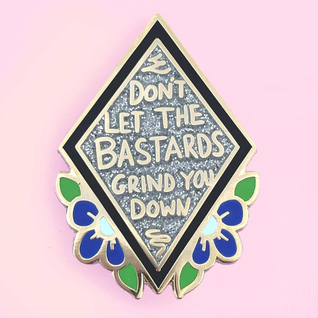Diamond shaped enamel lapel pin with a black outline, with the words 'Don't let the bastards grind you down' on a glittery silver background set inside the diamond shape. The pin is set against a pink background.