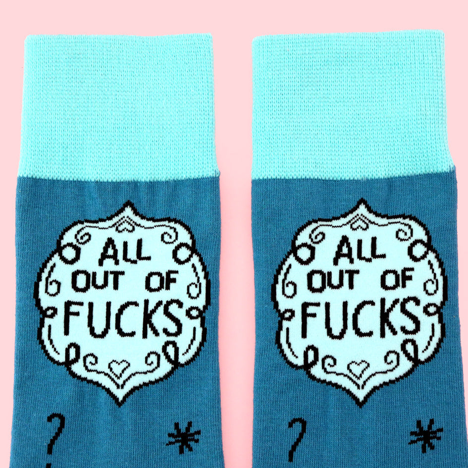 A pair of socks laying flat against a pink background. The socks are blue and light blue and read All Out Of Fucks.