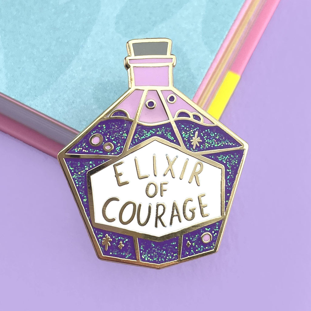 A hard enamel pin on a purple background. The pin is in the shape of a bottle with purple glitter. The pin reads Elixir Of Courage.