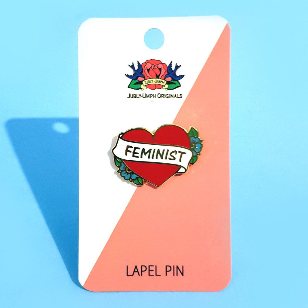 A hard enamel pin on Jubly-Umph backing card. The pin is in the shape of a red heart and reads Feminist.