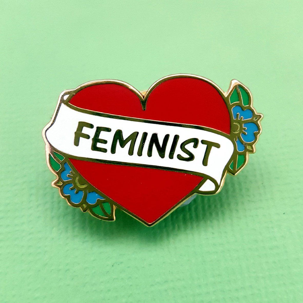 A hard enamel pin on a green background. The pin is in the shape of a red heart and reads Feminist.