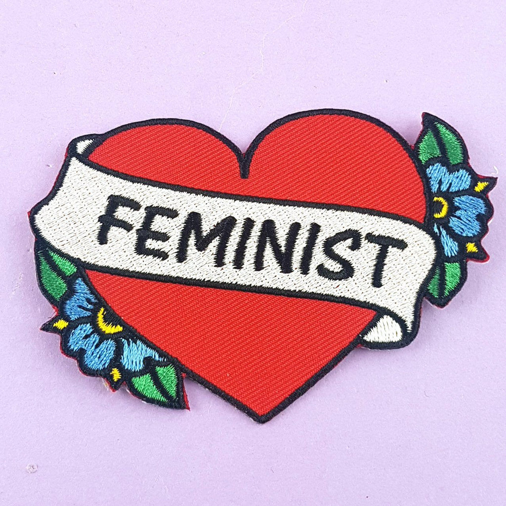 An iron on embroidered patch against a purple background. The patch is in the shape of a red heart and reads Feminist.