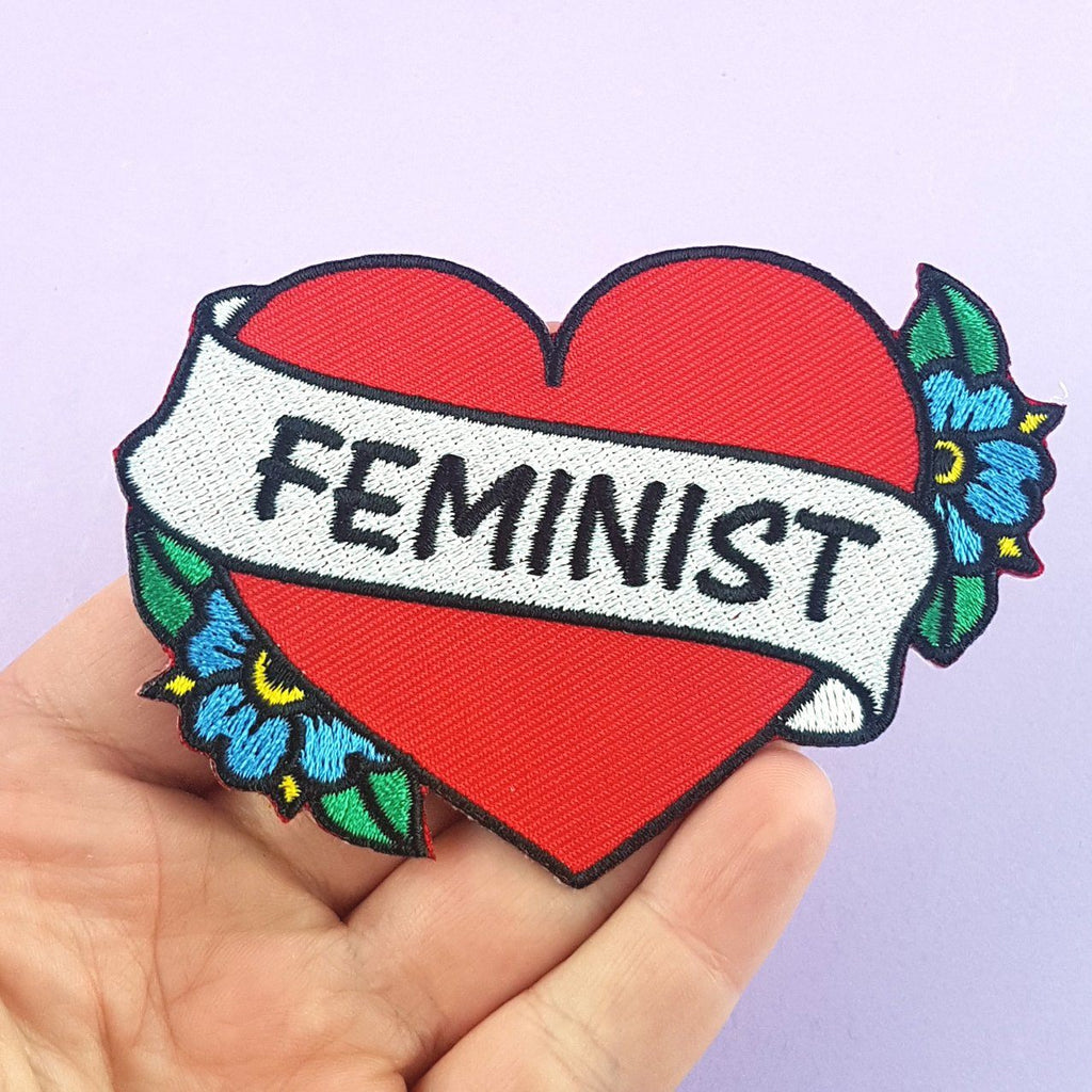 An iron on embroidered patch held in hand against a purple background. The patch is in the shape of a red heart and reads Feminist.
