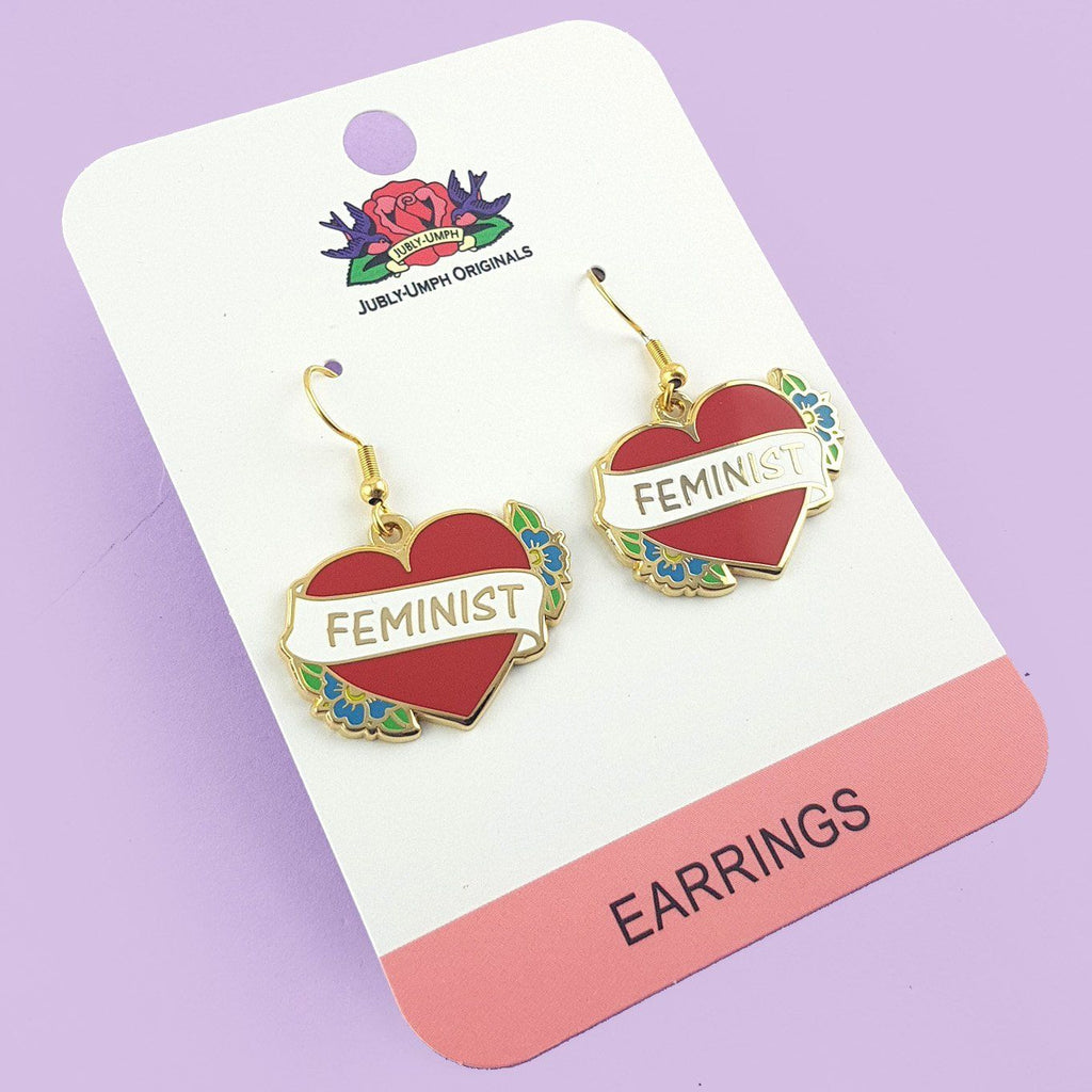 A pair of earrings displayed on Jubly-Umph card stock. The earrings are in the shape of a red heart and read Feminist.