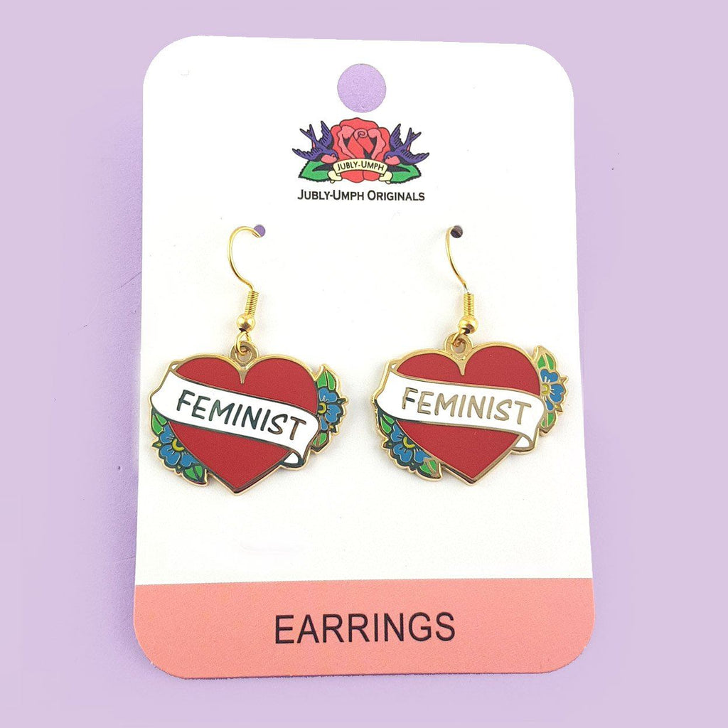 A pair of earrings displayed on Jubly-Umph card stock. The earrings are in the shape of a red heart and read Feminist.