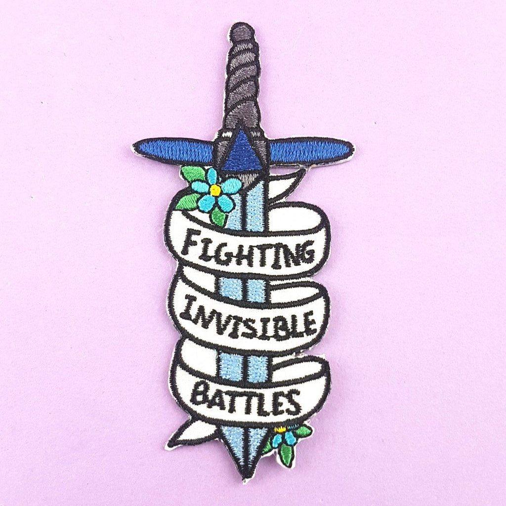 An iron on embroidered patch on Jubly-Umph against a purple background. The patch is in the shape of a dagger and reads Fighting Invisible Battles.