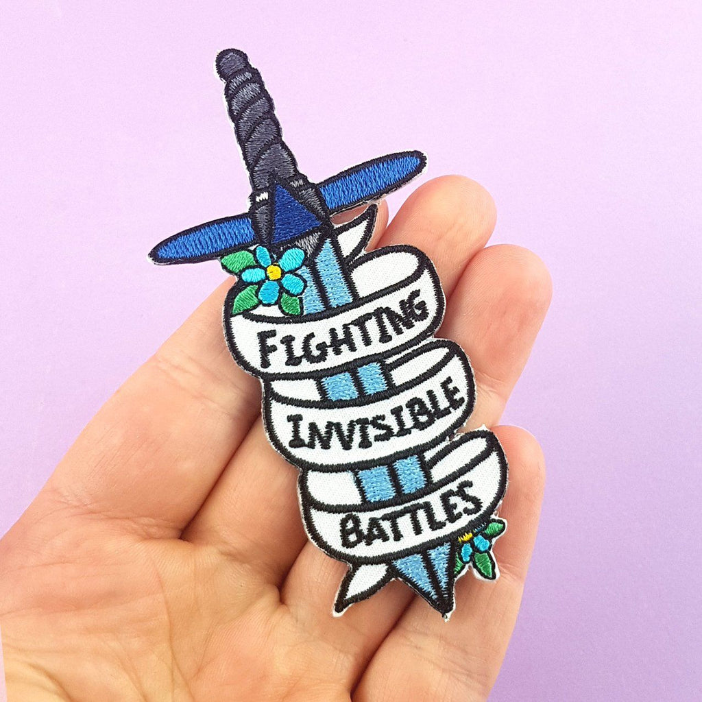 An iron on embroidered patch held in the hand against a purple background. The patch is in the shape of a dagger and reads Fighting Invisible Battles.