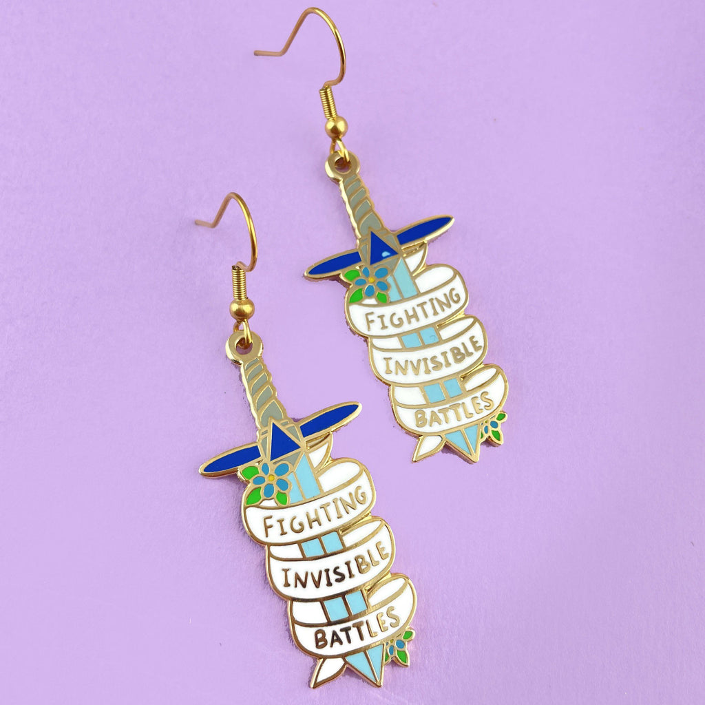A pair of earrings displayed on a purple background. The earrings are in the shape of dagger and read Fighting Invisible Battles.