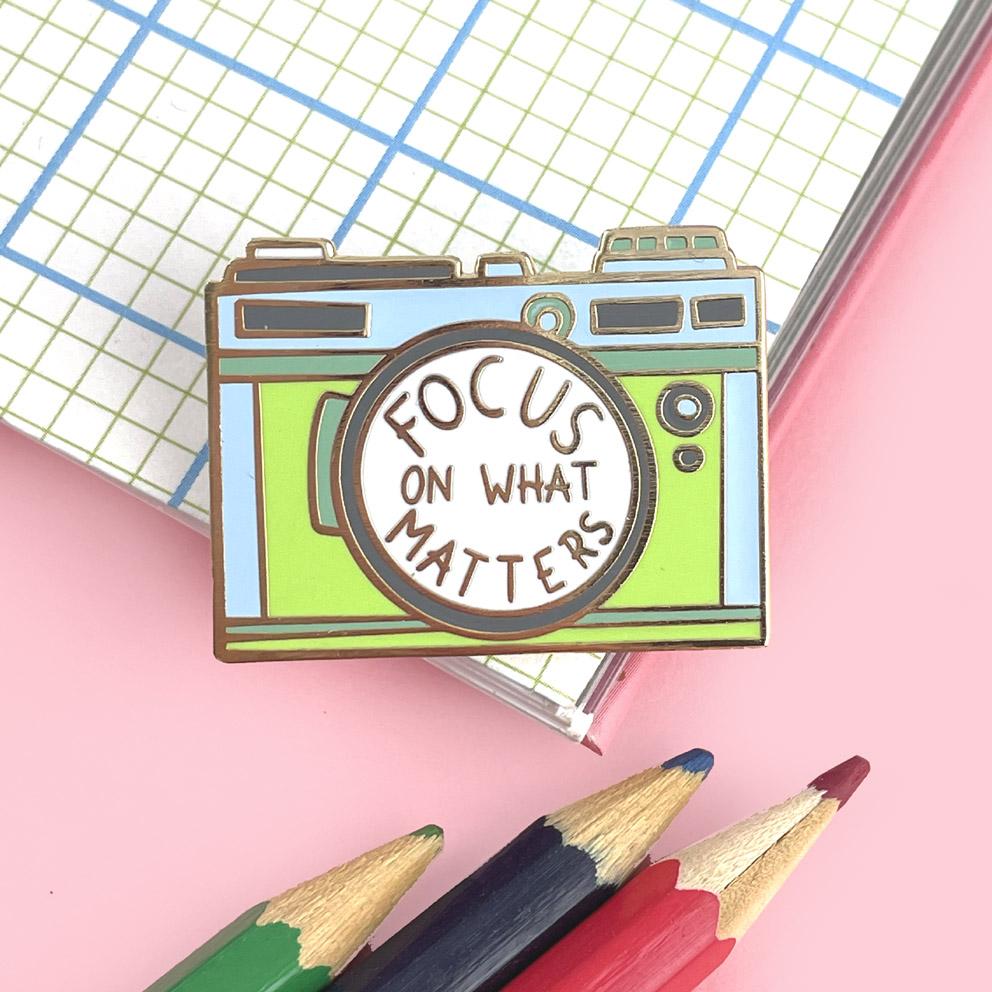 A hard enamel pin displayed with pencils. The pin is in the shape of a camera and reads Focus On What Matters.