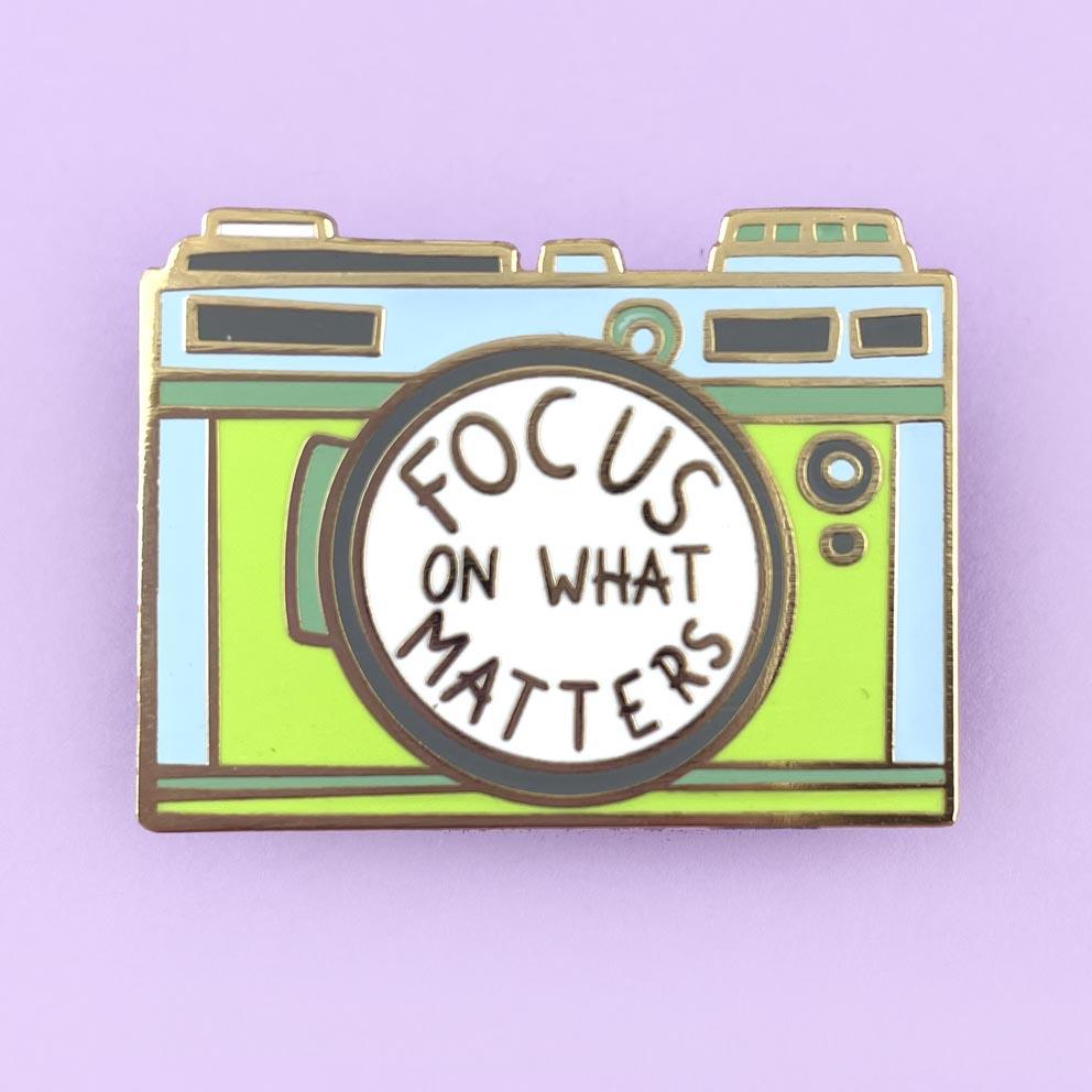 A hard enamel pin on a purple background. The pin is in the shape of a camera and reads Focus On What Matters.