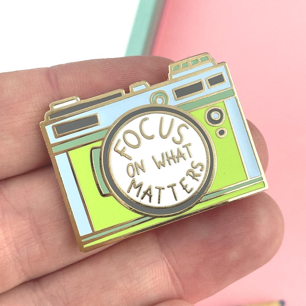 A hard enamel pin held in a hand. The pin is in the shape of a camera and reads Focus On What Matters.