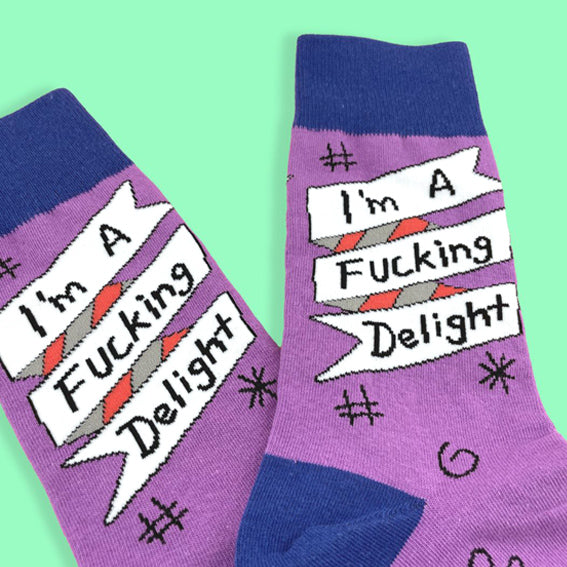 A pair of socks standing against a green background. The socks are purple and blue and read I'm A Fucking Delight.