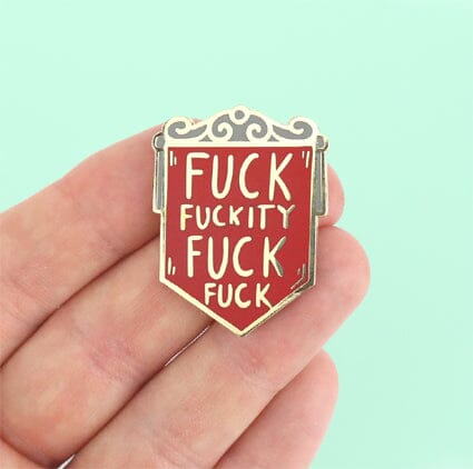 A hard enamel pin on held in the hand. The pin is in the shape of a red shield and reads Fuck Fuckity Fuck Fuck.