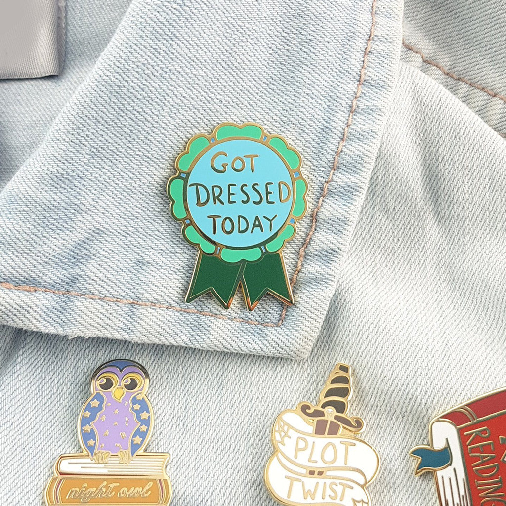 A hard enamel lapel pin displayed on a denim jacket with other Jubly-Umph lapel pins. The pin is in the shape of a green award ribbon. The pin reads Got Dressed Today.