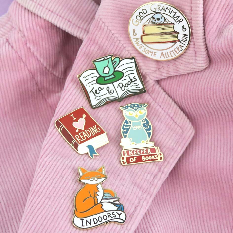 Hard enamel lapel pins on a pink jacket. The lapel pin is in the shape of a red book. The lapel pin reads I (heart image) reading. The other pins read Good Grammer & Awesome Alliteration, Tea & Books, Keeper of Books and Indoorsy.