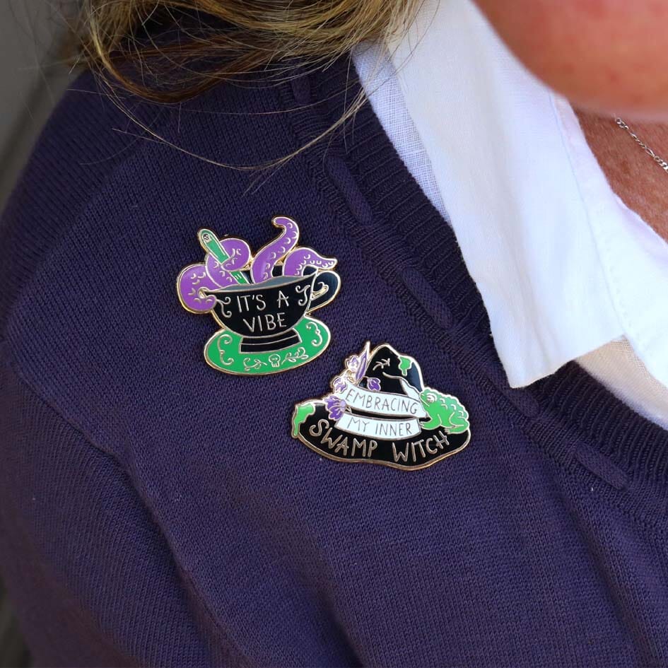 Two hard enamel lapel pin being worn on a purple shirt. The pin is black in the shape of a witches hat. The hat reads Embracing My Inner Swamp Witch. The second pin reads It's a Vibe.