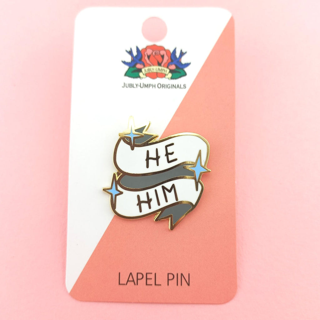 A hard enamel lapel pin on a Jubly-Umph backing card. The pin reads He Him.