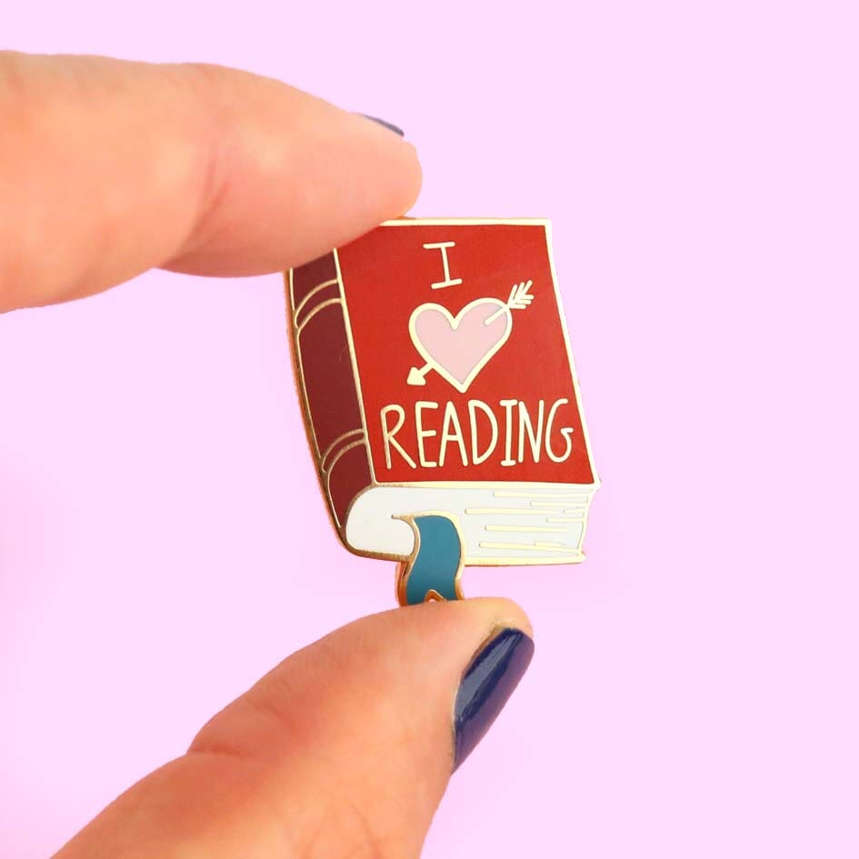 A hard enamel lapel pin held in a hand. The lapel pin is in the shape of a red book. The lapel pin reads I (heart image) reading.