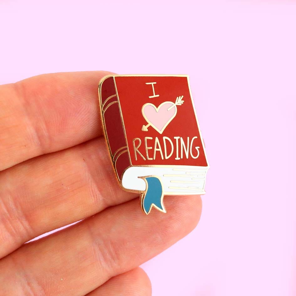 A hard enamel lapel pin held in a hand. The lapel pin is in the shape of a red book. The lapel pin reads I (heart image) reading.