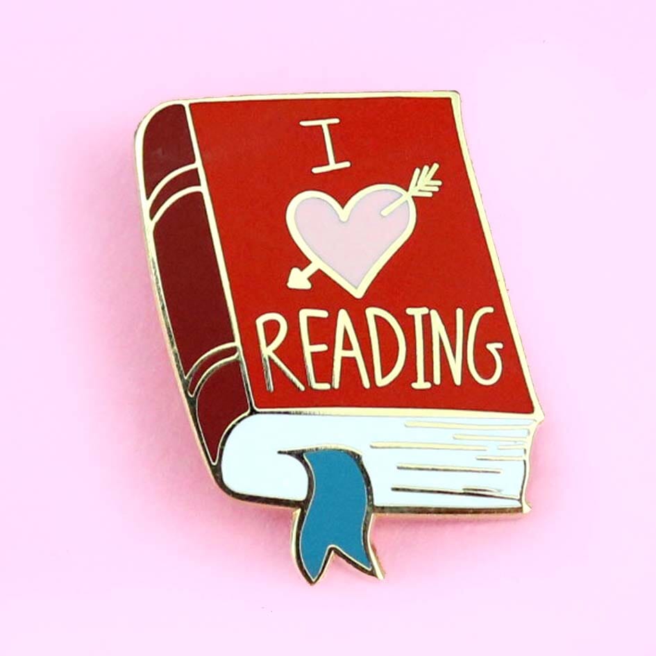 A hard enamel lapel pin displayed on a pink background. The lapel pin is in the shape of a red book. The lapel pin reads I (heart image) reading.