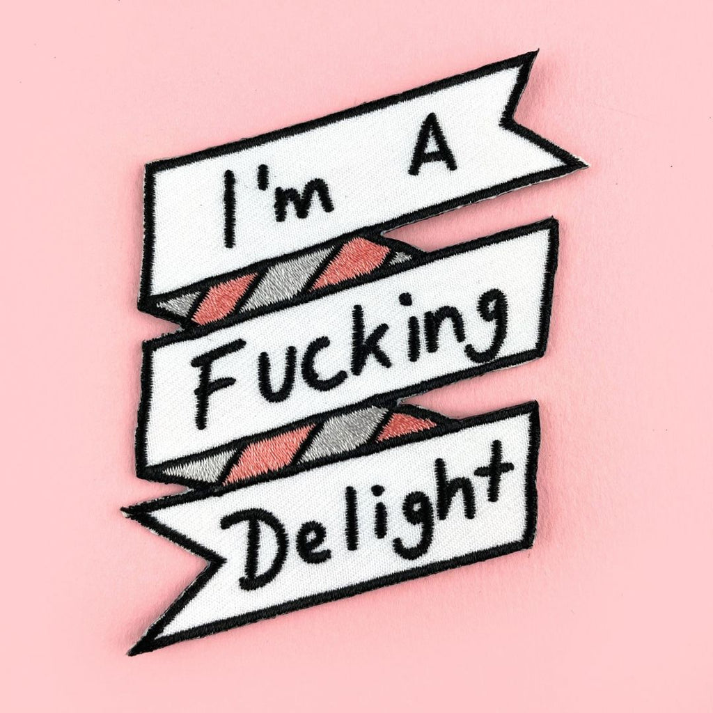 An iron on embroidered patch is being displayed on a pink background. The patch reads I'm A Fucking Delight.