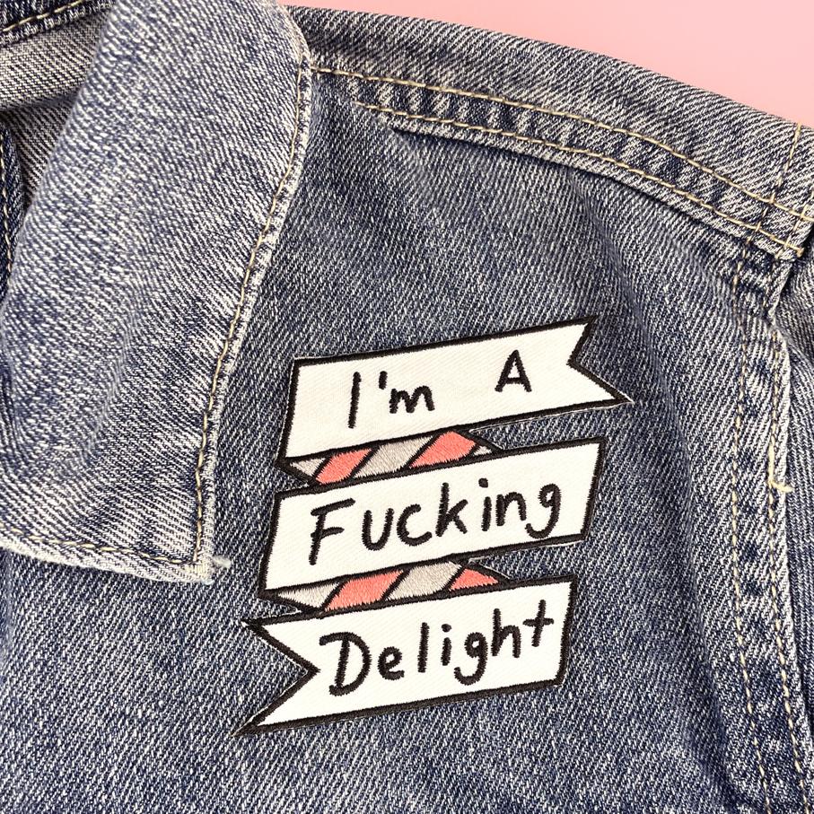 An iron on embroidered patch is being worn on a denim jacket. The patch reads I'm A Fucking Delight.