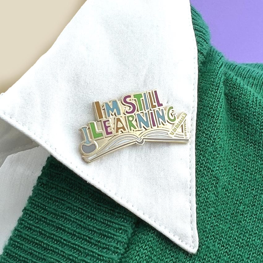A hard enamel lapel pin being displayed on a white collar. The lapel pin is an open book and reads I'm Still Learning.