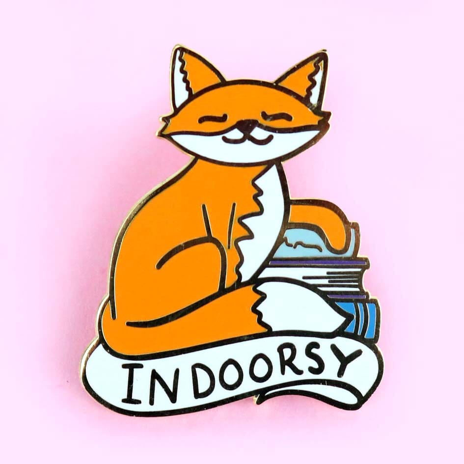 A hard enamel lapel pin displayed on a pink background. The lapel pin is in the shape of an orange cat and reads Indoorsy.