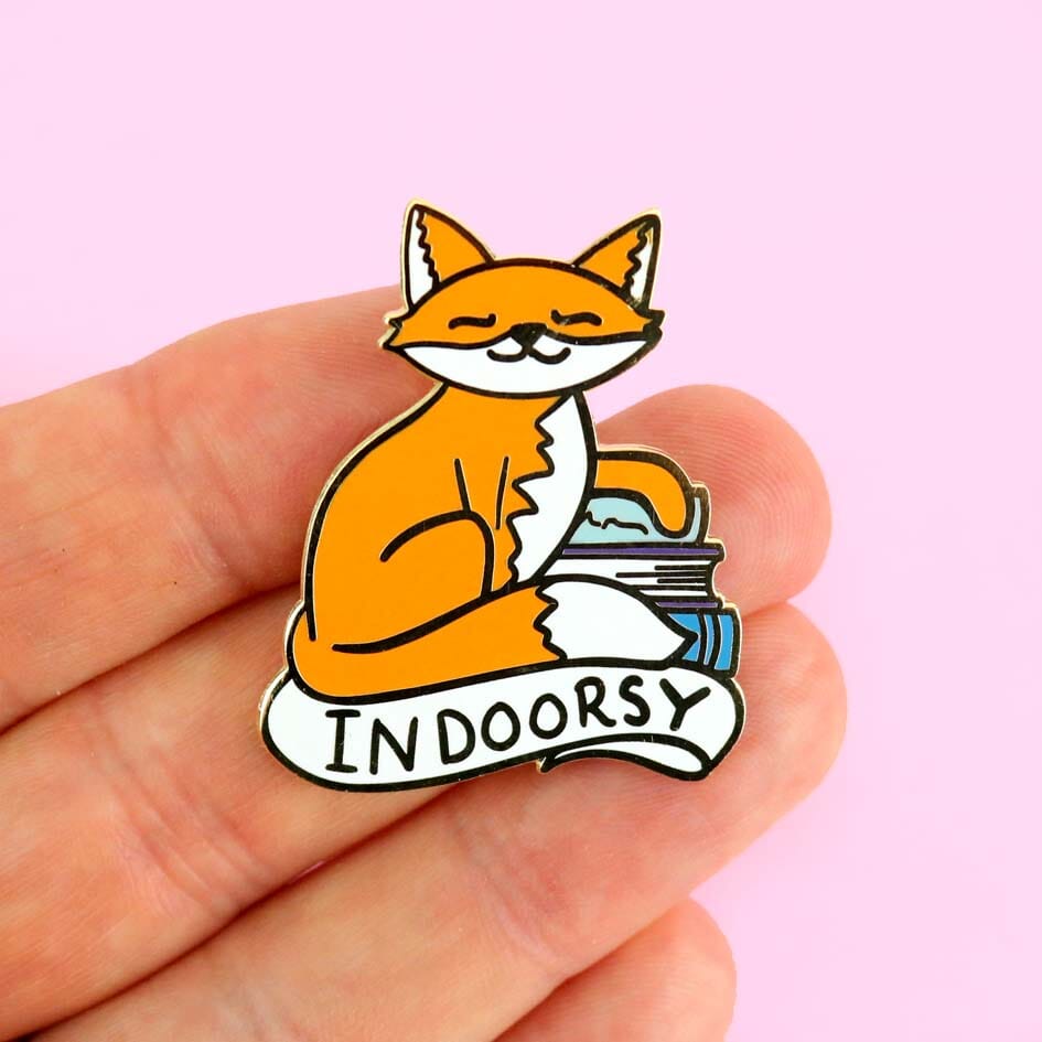 A hard enamel lapel pin being held in a hand. The lapel pin is in the shape of an orange cat and reads Indoorsy.