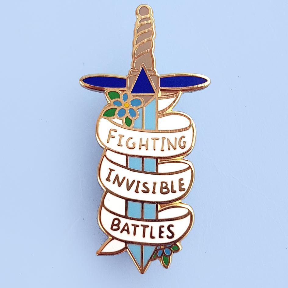 A hard enamel pin on a blue background. The pin is in the shape of a dagger and reads Fighting Invisible Battles.