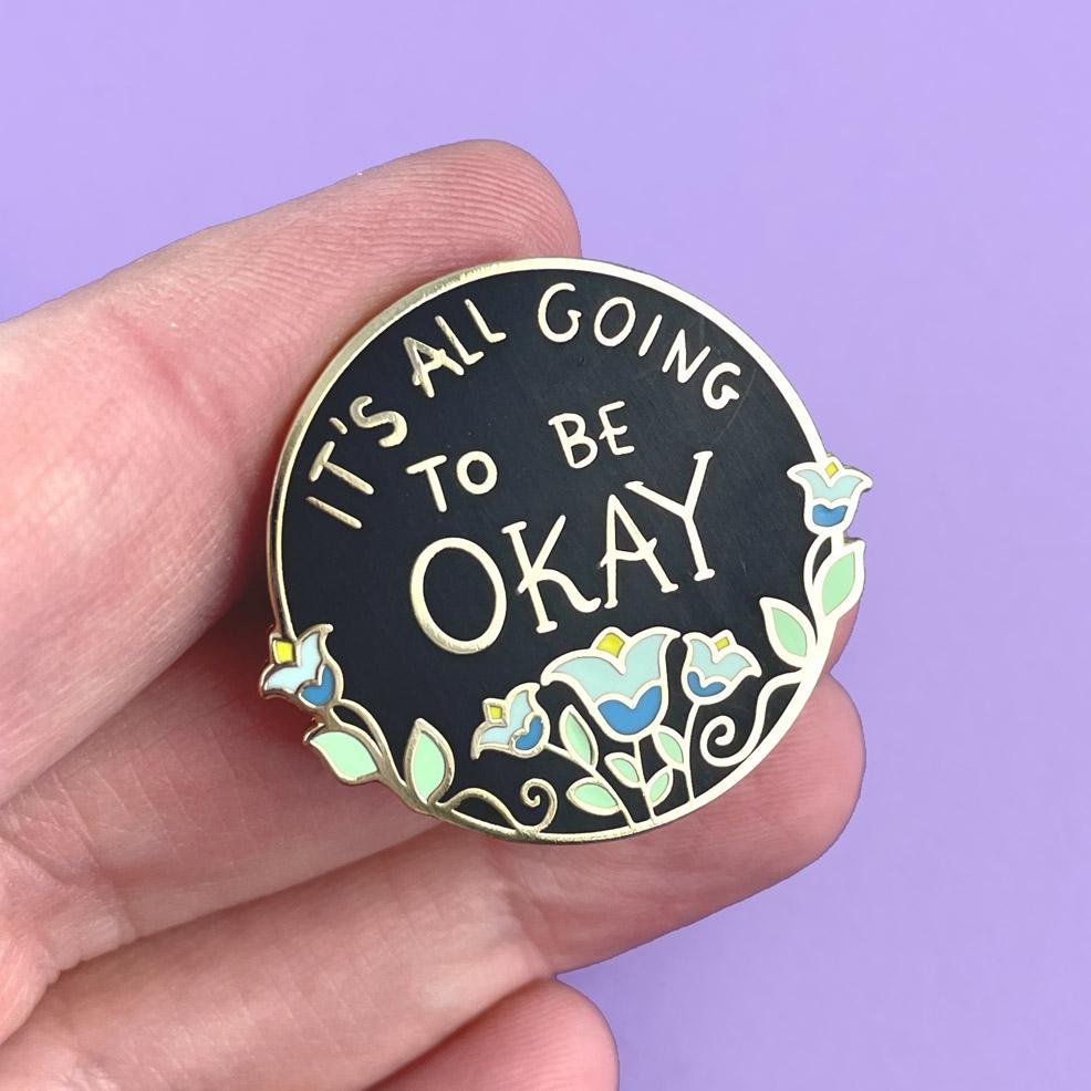 A hard enamel lapel pin being held in a hand. The pin is black with blue flowers. The pin reads It's All Going To Be Okay.