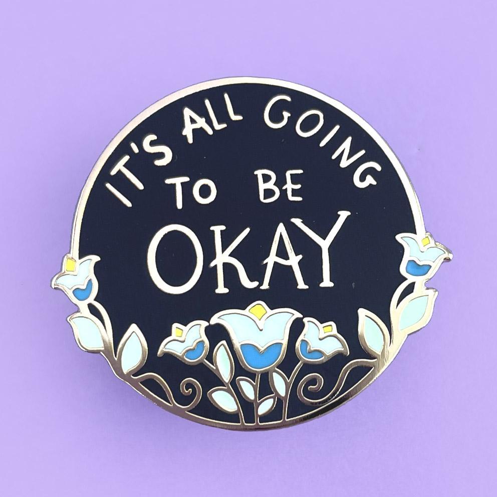 A hard enamel lapel pin displayed on a purple background. The pin is black with blue flowers. The pin reads It's All Going To Be Okay.