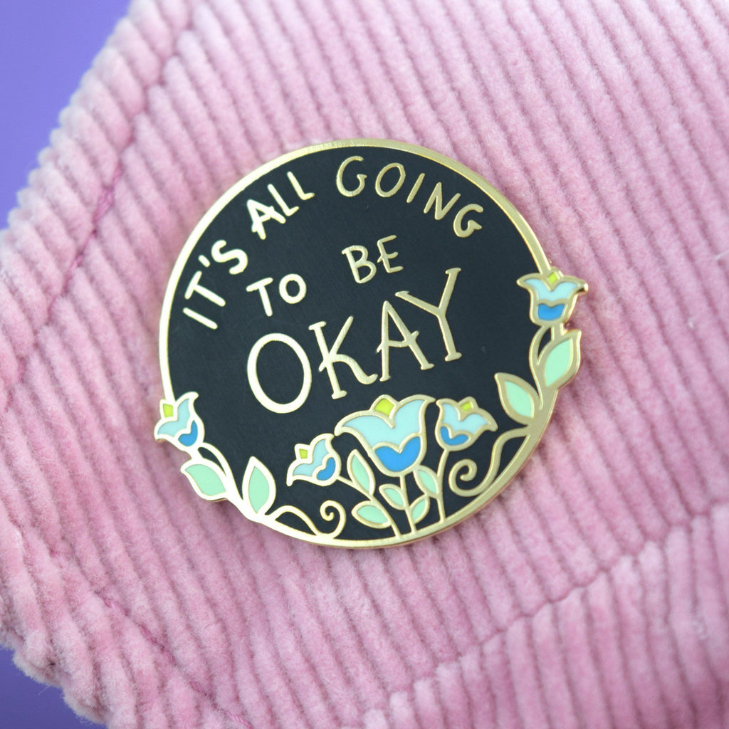 A hard enamel lapel pin displayed on a pink jacket. The pin is black with blue flowers. The pin reads It's All Going To Be Okay.
