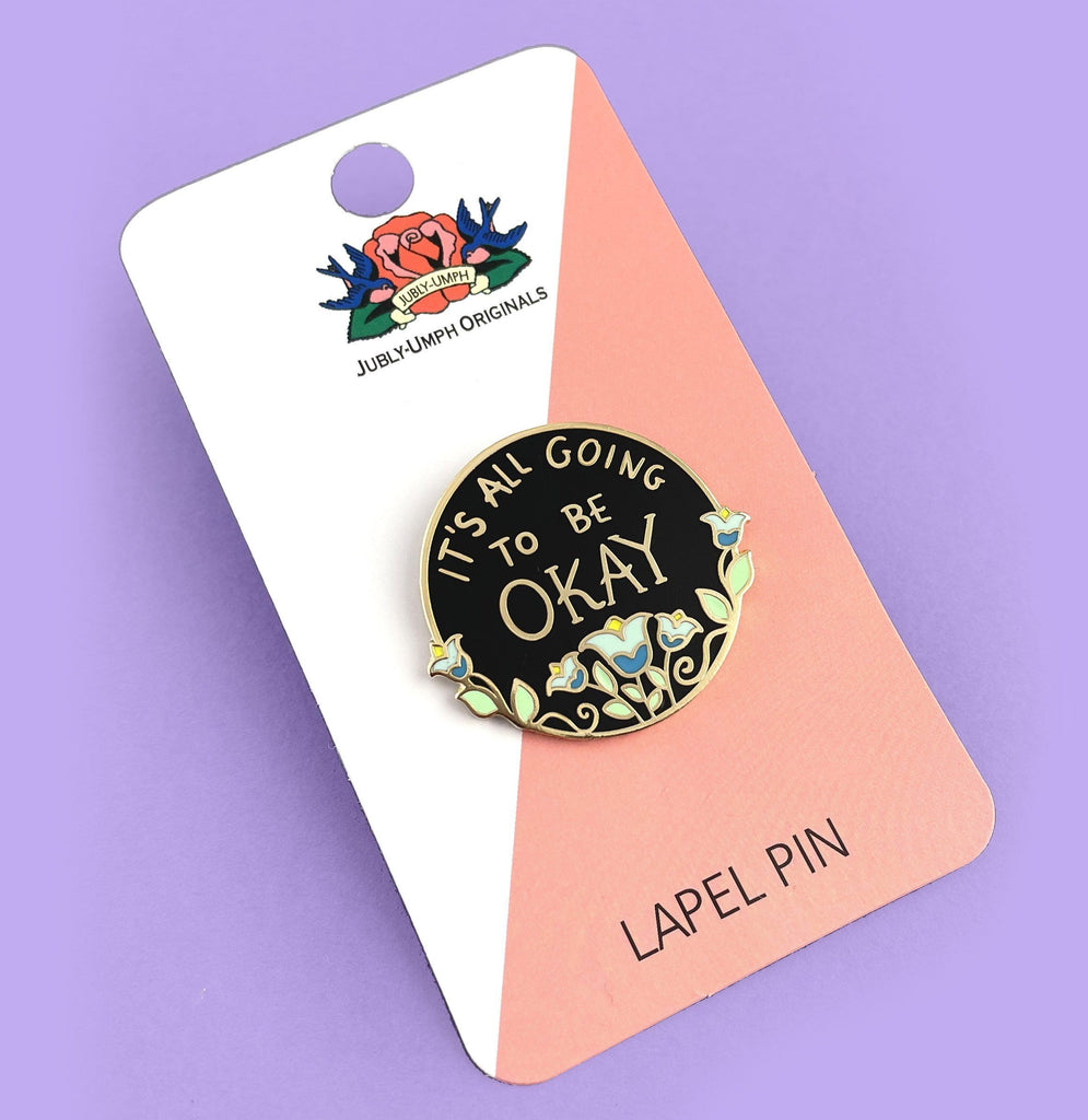 A hard enamel lapel pin displayed on a Jubly-Umph backing card. The pin is black with blue flowers. The pin reads It's All Going To Be Okay.