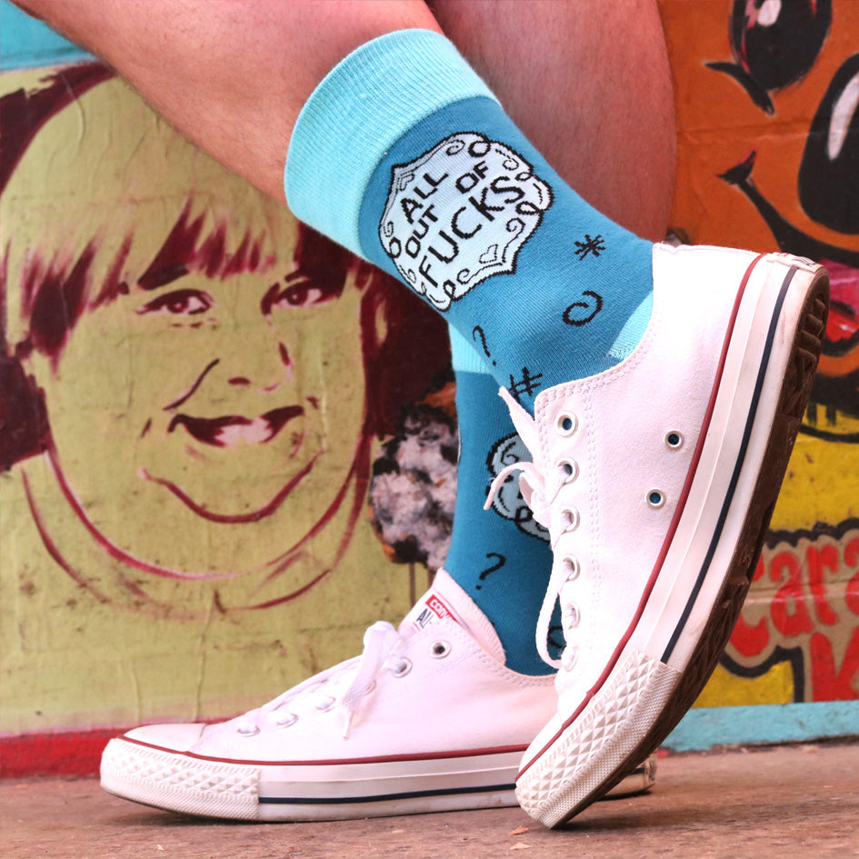 A pair of socks being worn with white shoes standing against a mural background. The socks are blue and light blue and read All Out Of Fucks.